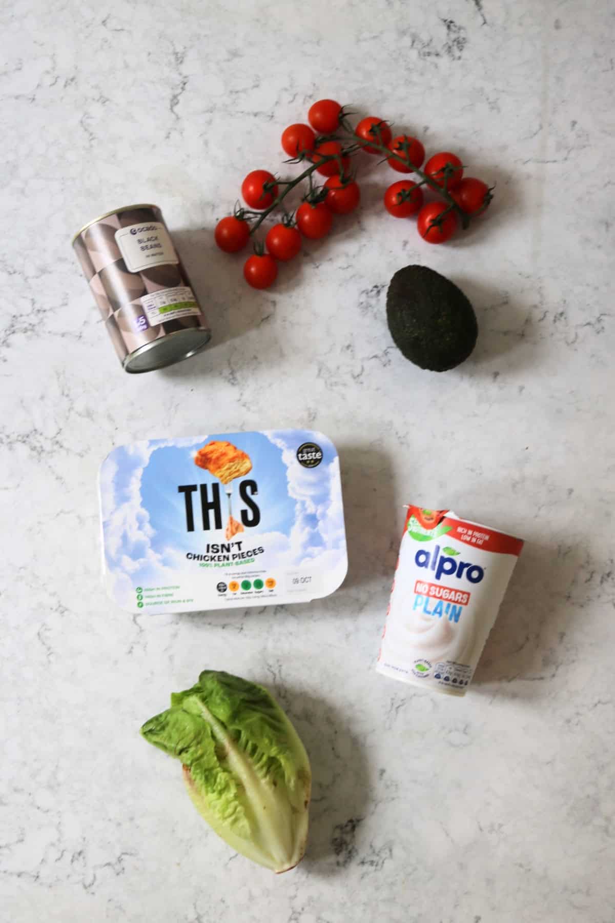 Cherry tomatoes, a tin of black beans, an avocado, a packet of vegan chicken pieces, vegan yogurt and a little gem lettuce laid out on a white marble counter top