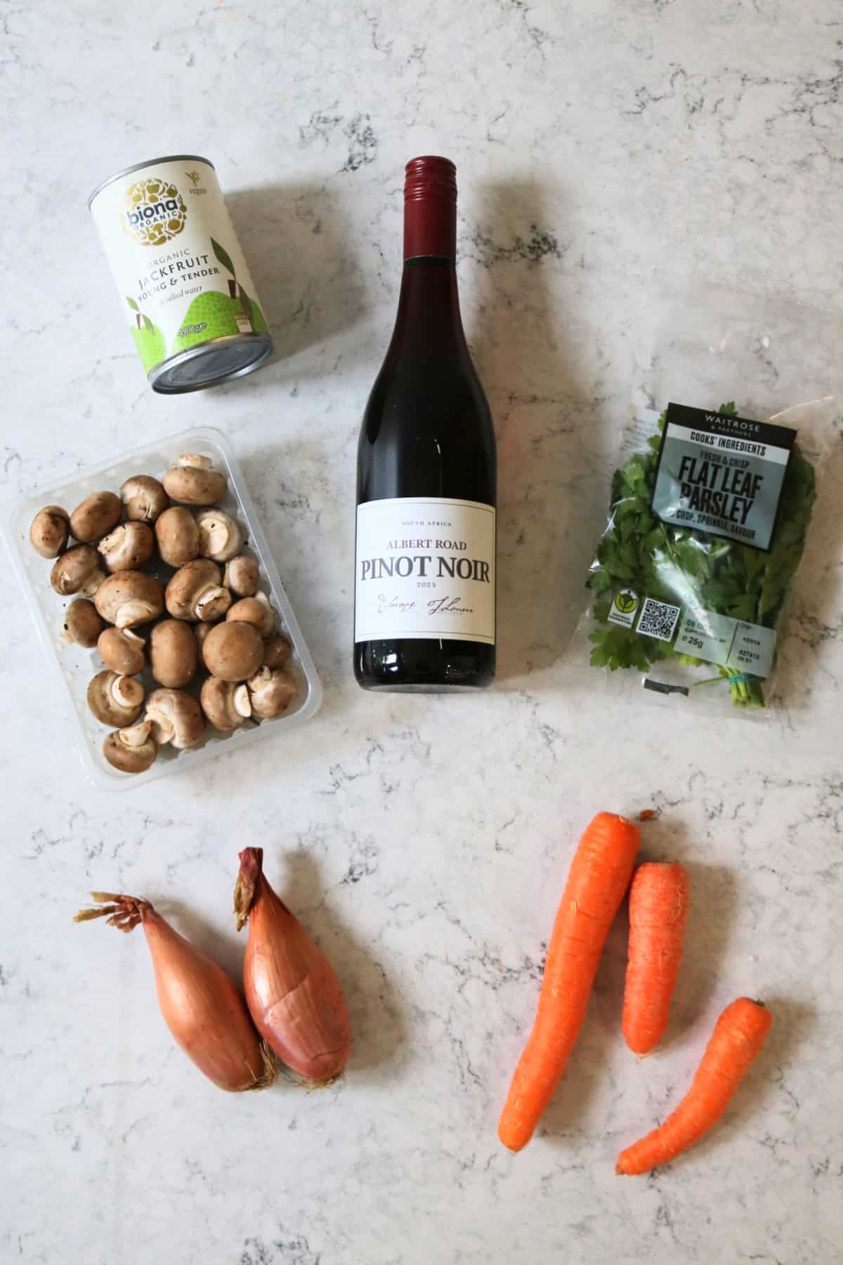 A tin of jackfruit, bottle of wine, chestnut mushrooms, flat leaf parsley, banana shallots and carrots laid out on a white marble counter top.