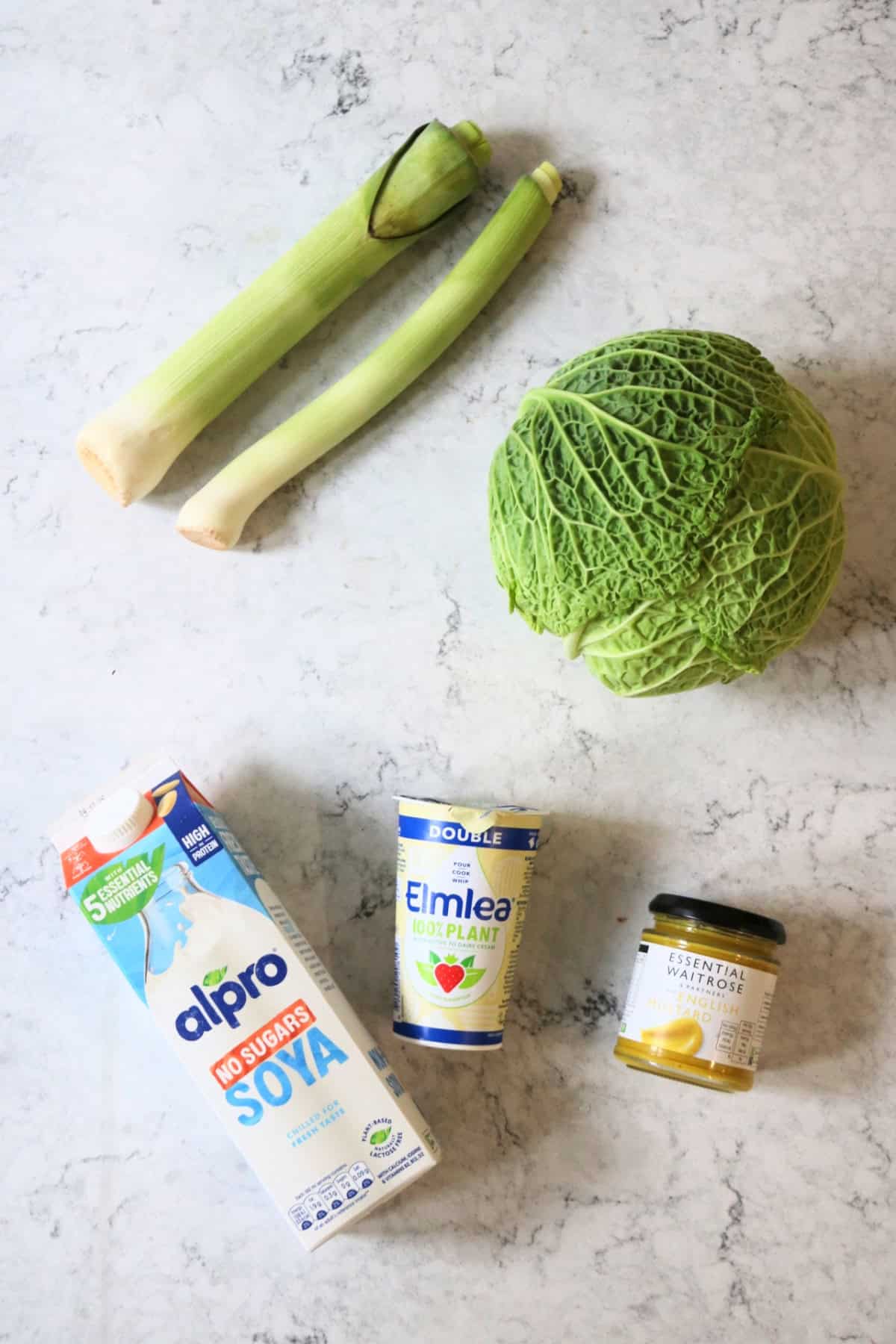 Two leeks, a head of cabbage, soy milk, vegan double cream and a jar of English mustard, all laid out on a white marble counter top