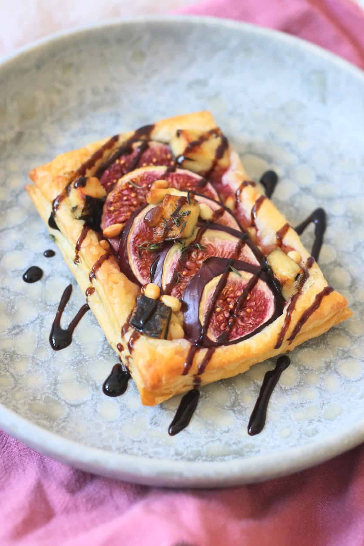 A Vegan Fig and Blue Cheese Tart on a blue plate, drizzled with balsamic glaze and served with a pink napkin