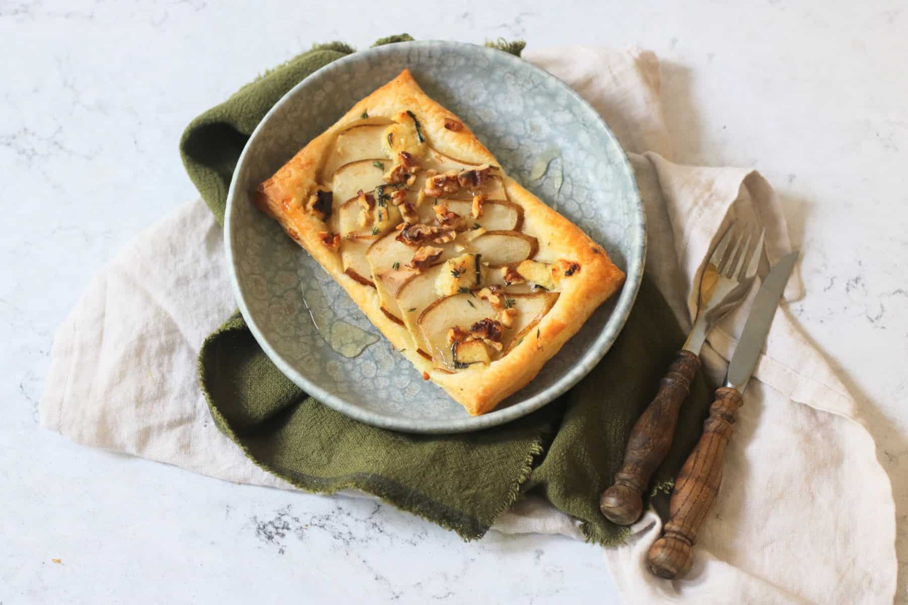 A Vegan pear and blue cheese puff pastry tart topped with walnuts, fresh thyme and vegan honey served alongside a green napkin and wooden handled cutlery