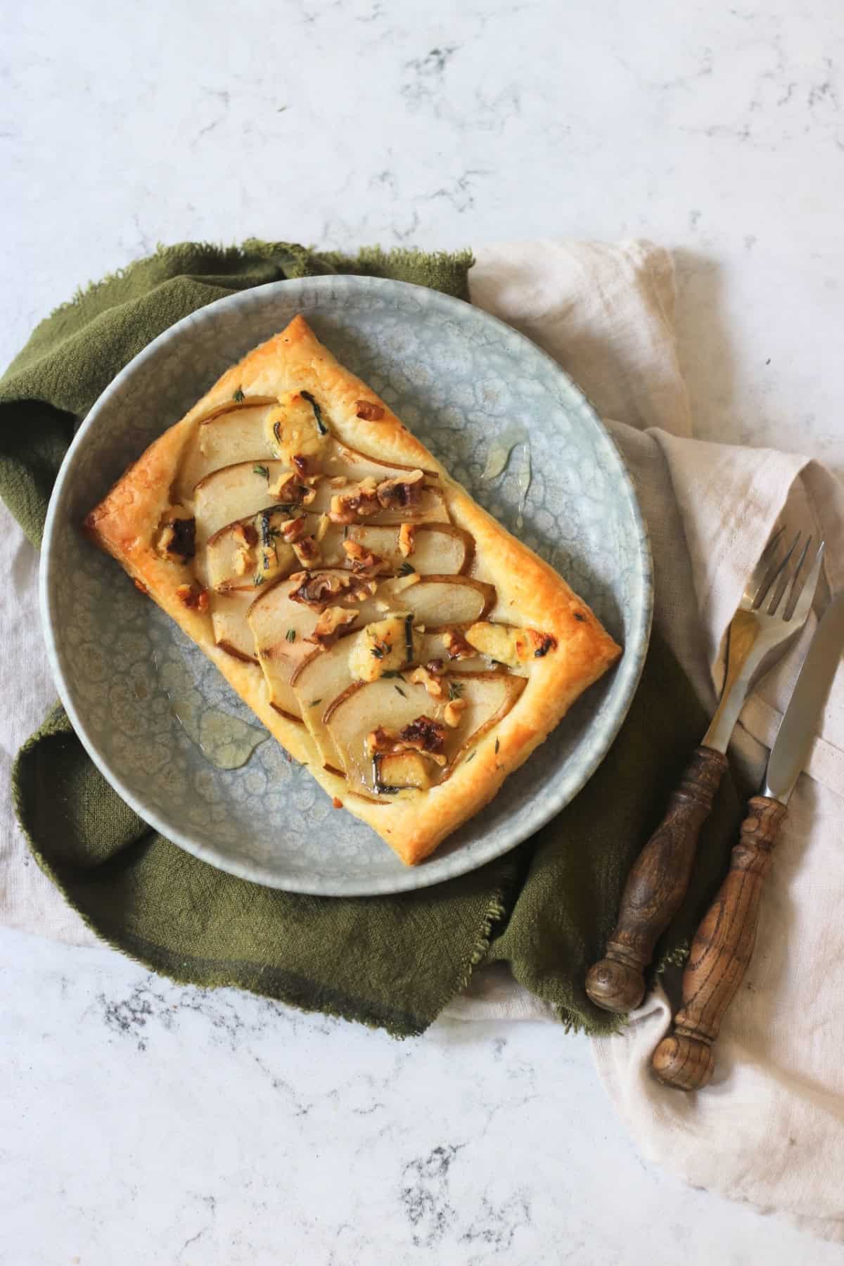 A Vegan pear and blue cheese puff pastry tart topped with walnuts, fresh thyme and vegan honey served alongside a green napkin and wooden handled cutlery