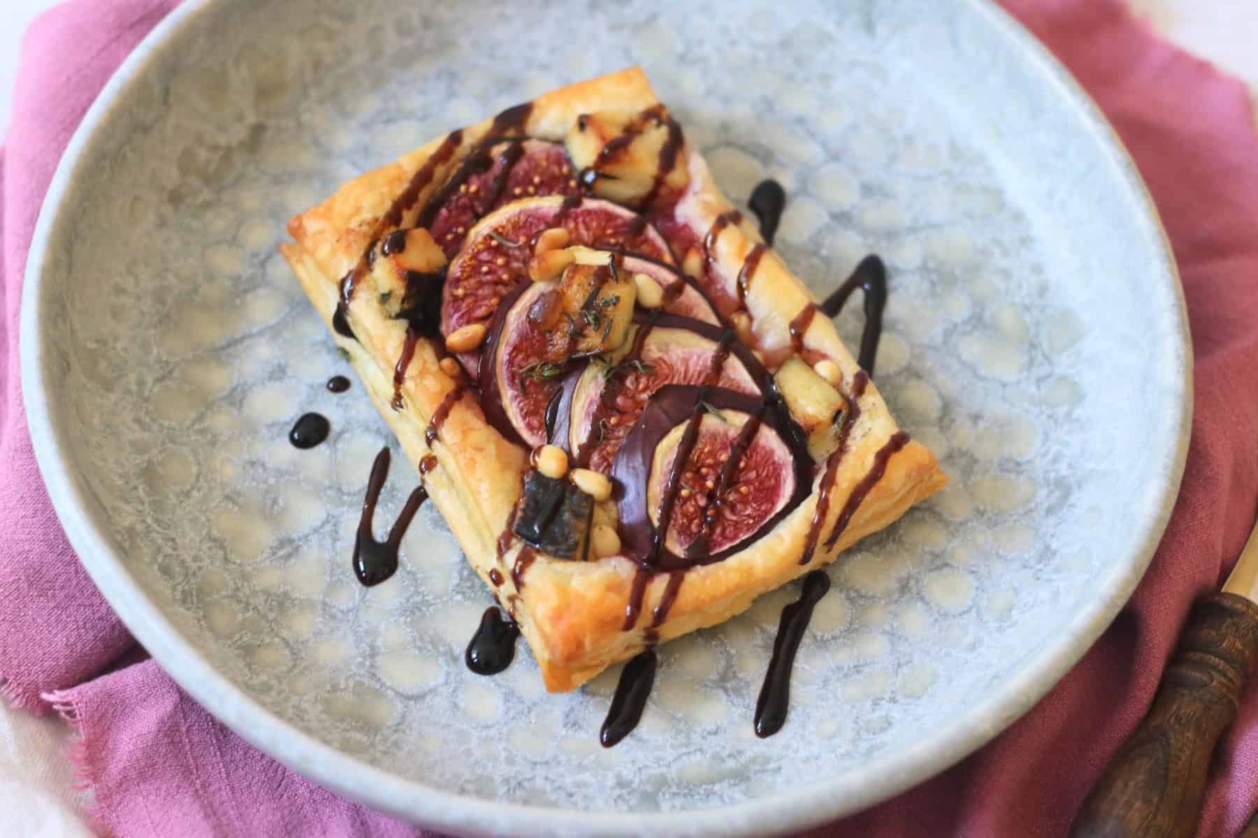A Vegan Fig and Blue Cheese Tart on a blue plate, drizzled with balsamic glaze and served with a pink napkin and wooden handled cutlery