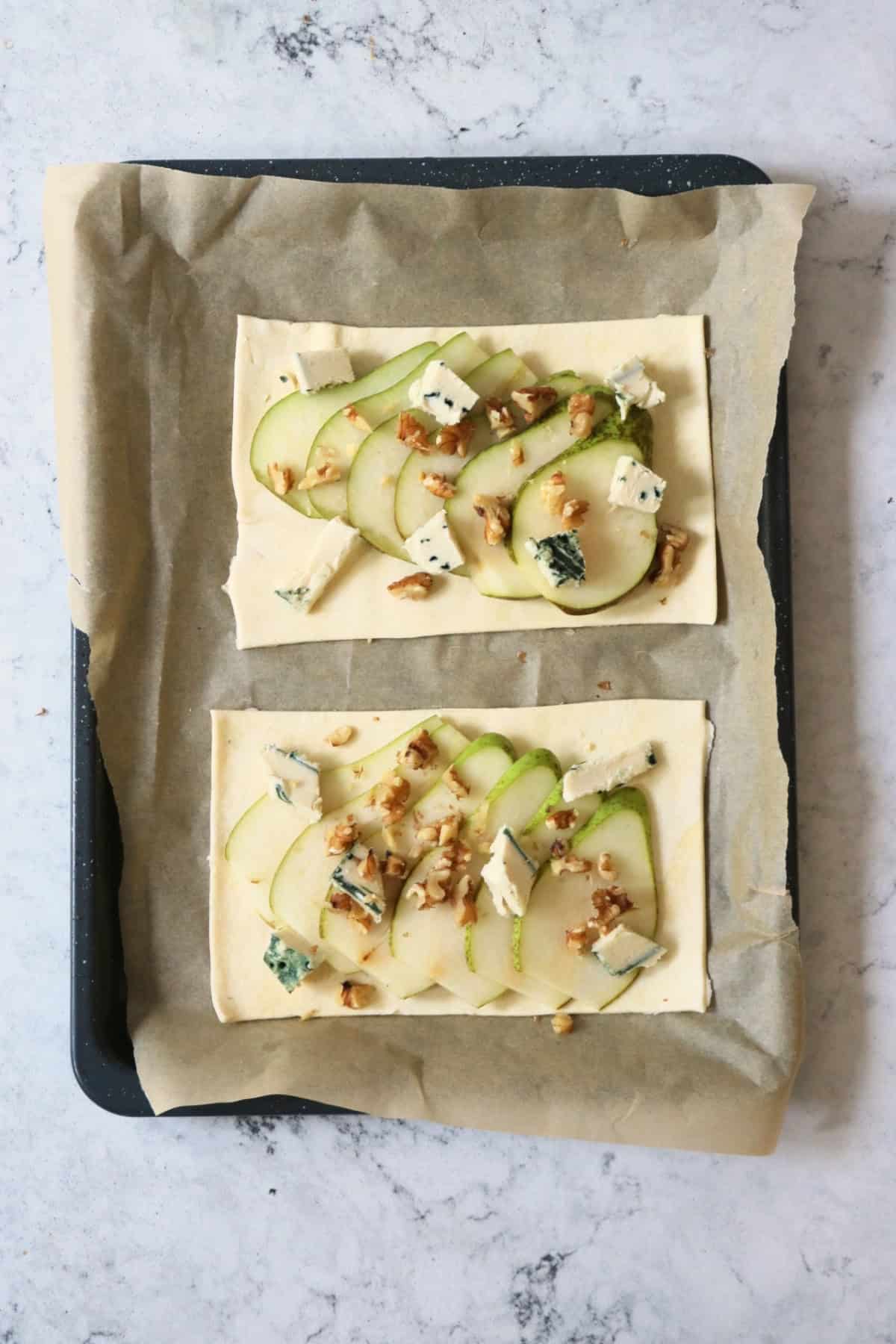 Two unbaked rectangles of puff pastry topped with sliced pear, crumbled vegan blue cheese and walnuts