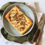 A Vegan pear and blue cheese puff pastry tart topped with walnuts, fresh thyme and vegan honey served alongside a green napkin and wooden handled cutlery