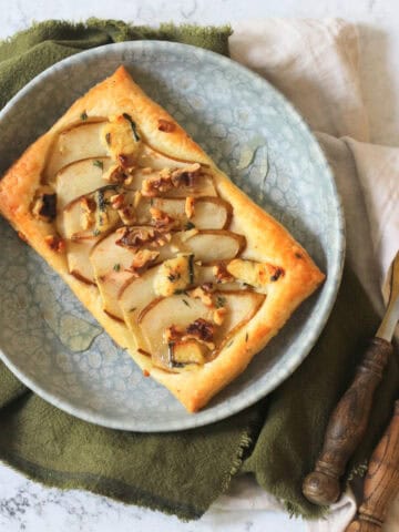 A Vegan pear and blue cheese puff pastry tart topped with walnuts, fresh thyme and vegan honey served alongside a green napkin and wooden handled cutlery