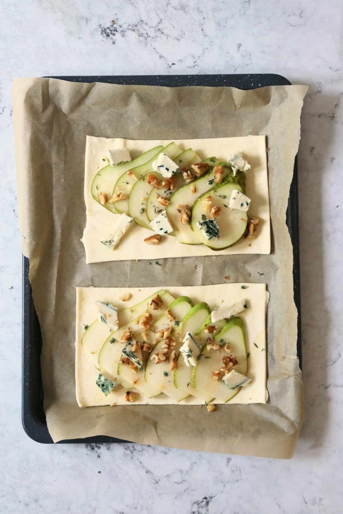 Two Vegan Pear and Blue Cheese Tarts topped with walnuts and fresh thyme on a baking sheet ready for the oven